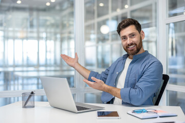 Fototapeta premium Young bearded man sitting at a desk with a laptop, smiling, and gesturing with open hands, presenting or welcoming in a bright, modern office workspace