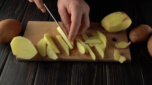 Hands slicing fresh potatoes into thin strips on a wooden cutting board with whole potatoes visible on a rustic wooden table