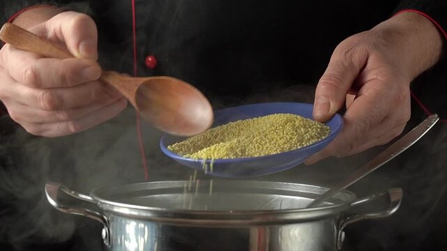 Chef pours couscous from a blue bowl into a steaming pot using a wooden spoon, showcasing the cooking process in a kitchen setting