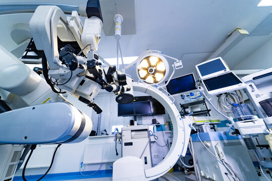 Surgical microscope and mobile C-arm imaging system in neurosurgery room. Profile view of neurosurgical microscope with C-arm fluoroscope and medical monitors in the background.