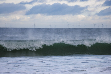 Spray from large breaking sea wave in front of offshore wind farm turbines