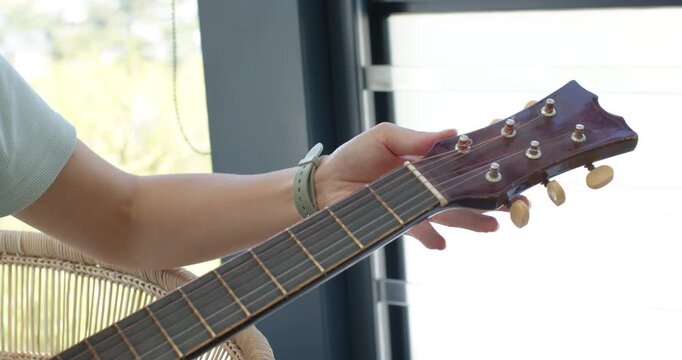 Left hand is touching tuning pegs and tightening strings to tune acoustic guitar by window