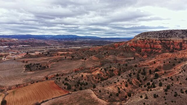 Wide view of a canyon with red rock shapes and patches of grass. Clouds cover the sky. The land is rocky with some flat areas. Hills rise in the background, adding depth to the scene.