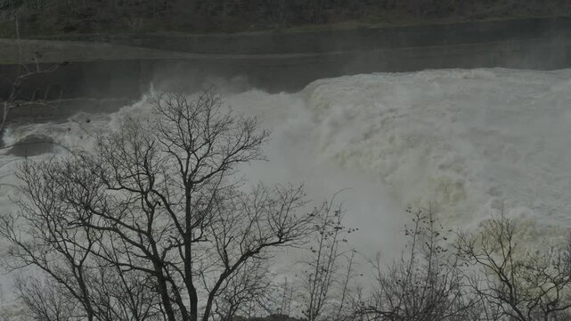 Massive Water Discharge from Dam with Nature Elements in Foreground. A brutal and powerful cascade of white rushing water discharging from a dam spillway. Close-up shot featuring tree branches 