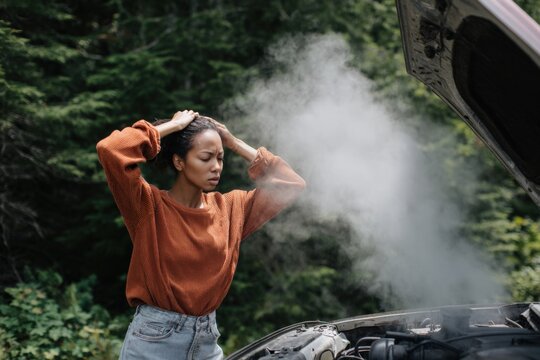 Frustrated young african female examining overheated car engine in forested area
