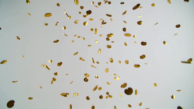 Releasing from upper frame, cloud of gold confetti descending and rotating over pale gray backdrop