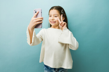 Tween girl with long hair taking a selfie on a smartphone, showing peace sign and smiling for...