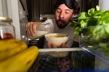 Latin man wearing sleep mask pouring milk into cereal bowl late at night from inside open refrigerator