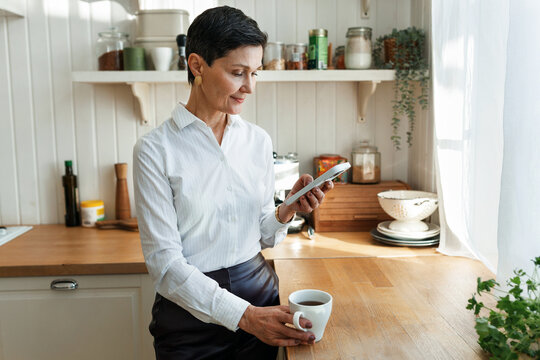 Side view of mature elegant lady in office clothes having coffee at home kitchen using smartphone before going to work, browsing news feed, chatting in chat bot via mobile applications