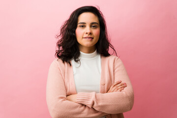 Obraz premium Hispanic woman biting her lip with crossed arms, expressing thoughtfulness and self-assurance on a pink background
