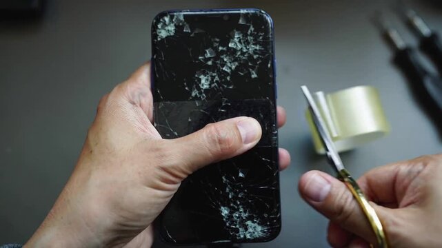 Placing phone upright, adult hands pulling and trimming tape with scissors on bench to secure glass
