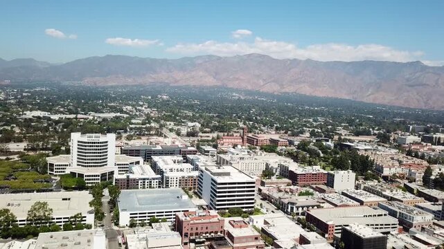 Aerial view of downtown Pasadena, California, showing city buildings, residential blocks, tree-lined streets, and the San Gabriel Mountains in the background under clear daylight