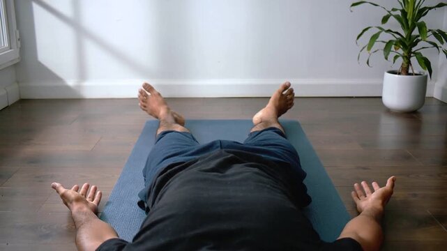 Pulling back camera revealing man lying on blue mat, wearing black tee, near potted plant resting