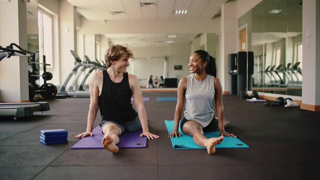 Making eye contact, workout partners in athletic wear stretching hamstrings on yoga mats, chatting