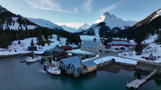 Approaching drone revealing blue boathouses on pilings at snowy coastal harbor, showing smoke plume