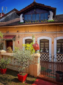 Street view in Panaji, India, with a terrace of red flower pots in the foreground and a beige colonial-era house with white trim and a mezzanine beyond, reflecting historic charm.