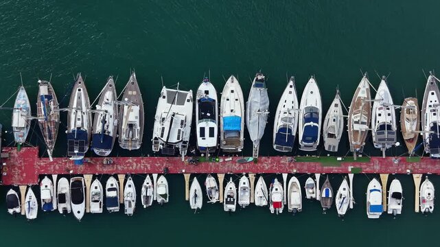 Boats are parked in rows at a marina with a red dock. The water is clear and reflects the blue sky. People are seen moving around on the dock and on some boats.