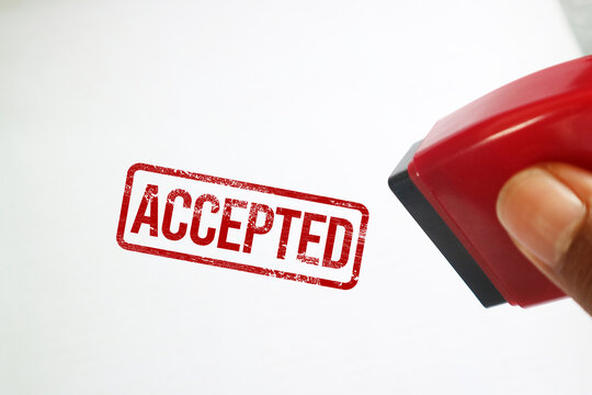 Hand holding a red rubber stamp next to a red ACCEPTED ink imprint on a clean white paper background. Conceptual image for approval.