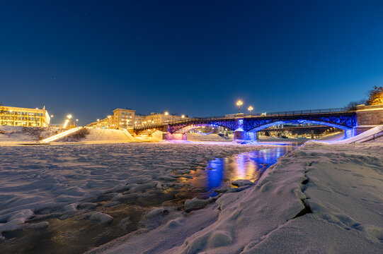 Night view of the frozen Neris River in Vilnius with Žvėrynas Bridge illuminated in red and blue lights, the Lithuanian Parliament in the background, and faint stars visible in the winter sky.