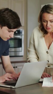 Vertical video: Typing teen, mom in cream cardi reviewing homework at kitchen counter with laptop