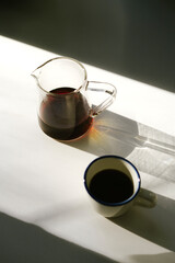 A cup of freshly brewed hot black filter coffee and a server full of it, standing on the white surface in the sun, making soft golden shades, top view, flat lay, vertical image 