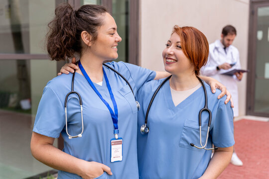 Two female nurses in blue scrubs, embraced and smiling, standing outdoors with a doctor in the background