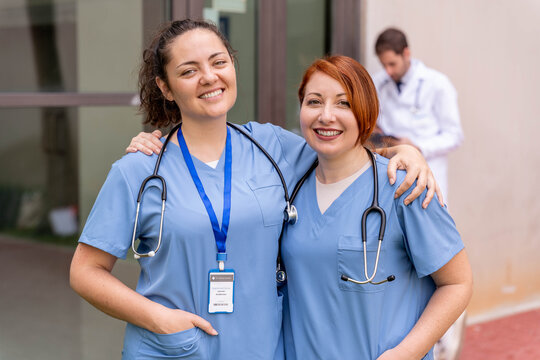 Healthcare team smiling, two happy nurses wearing scrubs and stethoscopes working at hospital, male doctor in background