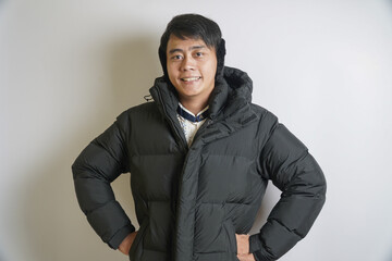 Smiling young Asian man confidently posing in a warm black puffer jacket and cozy earmuffs against a clean studio background, perfect for showcasing winter fashion and comfort © yashis007