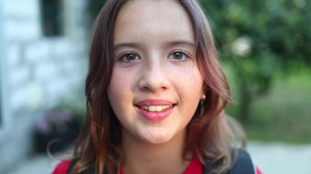 Girl in a red shirt smiles while standing outside on a sunny day, licking lips and enjoying the moment with friends nearby