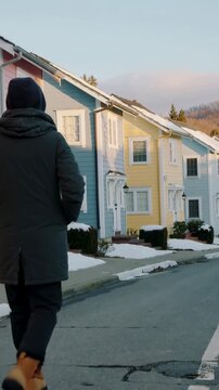Vertical video: Stepping man in dark parka tan boots walking sidewalk by pastel houses, copy space