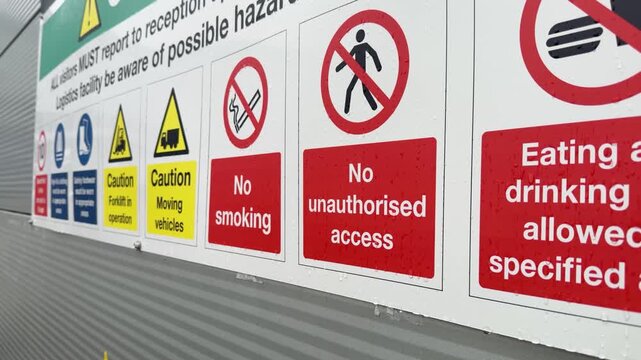 Collection of health and safety warning signs including no smoking and no unauthorised access displayed on a wet outdoor wall