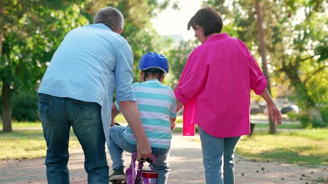 Family day out,dad mom teaching son ride bike in park. Child cyclist with parents in city park. Happy family, small child in helmet, with father mother, playing on bike outdoors in summer. People park