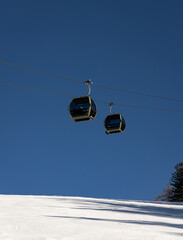 Two cable car cabins suspended above a snowy ski slope under a deep blue sky. Minimal winter mountain scene with modern gondola lift, clean composition and copy space.