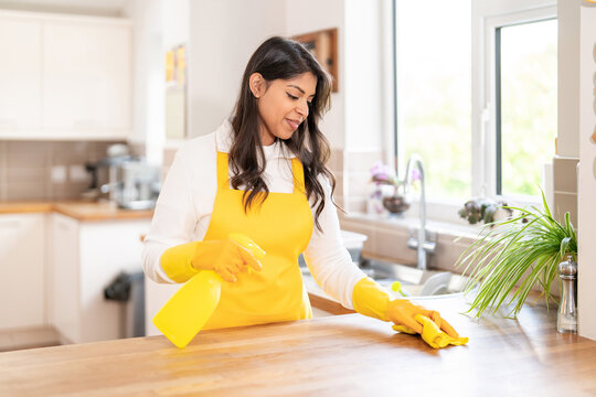 Woman cleans kitchen counter with spray and cloth while wearing yellow gloves and apron