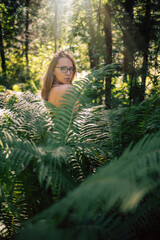Naklejka premium Back view of a woman standing in a lush green forest, touching her hair while sunlight filters through the trees and ferns.