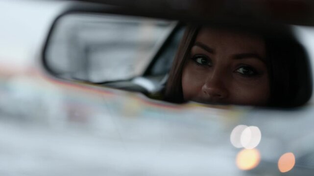 A stylish woman driver in her car turns the rearview mirror and locks eyes with the camera through the reflection. Soft focus emphasizes her confident, direct look.