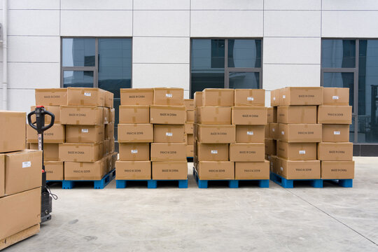 Stacked cardboard boxes on blue pallets with a hand pallet truck outside a warehouse building
