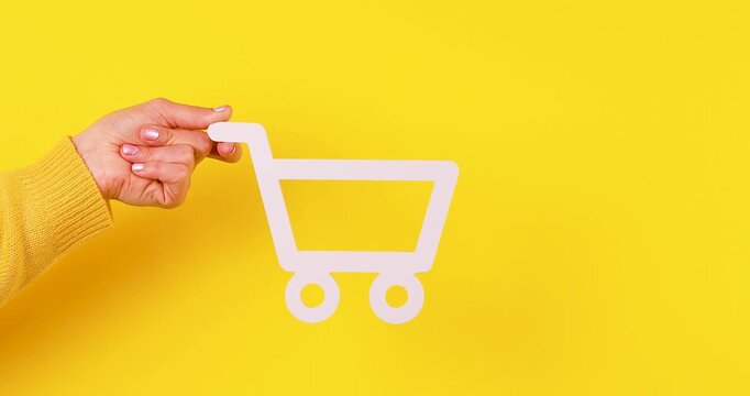 Close-up of a hand holding a shopping cart symbol on a bright yellow background. Concept of online shopping, e-commerce, retail, purchasing, and consumer behavior. Isolated, high contrast, 4K video.