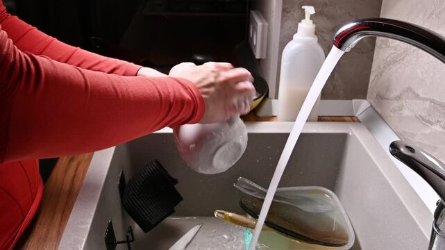 A close-up with localized lighting. The hands of a Caucasian woman washing dishes in a home kitchen late at night in the dark.