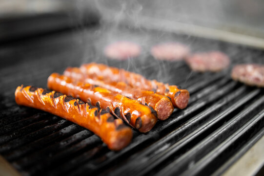 Grilled sausages with spiral cuts and char marks sizzle on a hot metal barbecue grate with rising smoke.