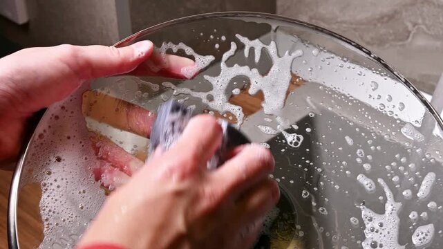A close-up with localized lighting. The hands of a Caucasian woman washing dishes in a home kitchen late at night in the dark.