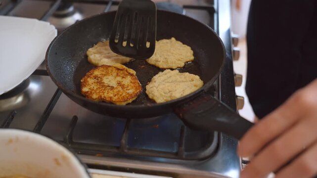Close-up view of a woman's hands using a spatula to fry traditional golden potato pancakes, or draniki, in hot oil on a gas stove, preparing a delicious homemade meal in the kitchen