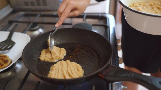 Close-up view of a woman's hands using a spatula to fry traditional golden potato pancakes, or draniki, in hot oil on a gas stove, preparing a delicious homemade meal in the kitchen