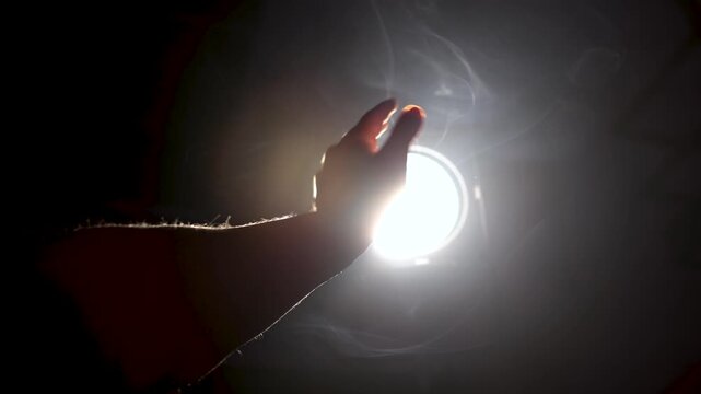 A man's hand stretches out towards a intense beam of light in a hazy atmosphere. Soft-focus close-up symbolizes the search for divine truth and revelation.