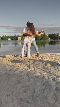 Romantic couple dancing bachata on sandy beach by lake at sunset. Man and woman moving together outdoors, enjoying summer evening light, music and love