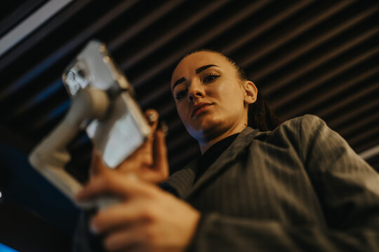 A confident woman looks down at a handheld device in a low-angle shot. She wears a striped jacket and stands indoors, focused on the screen.