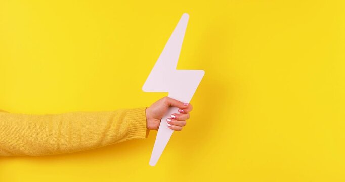 Close-up of a hand holding a lightning symbol simulating an electric shock on a bright yellow background. Concept of energy, power, electricity, danger, speed, and high voltage. Isolated, high contras
