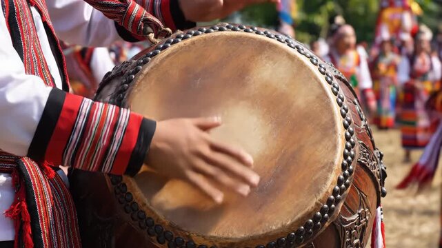 Close up of hands playing a large traditional drum outdoors during a festival with people in colorful native attire in the background under bright sunlight