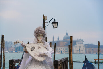 Elegant performer in white Venetian carnival costume playing a lute near gondolas on the lagoon in Venice, Italy