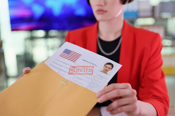 A woman in a bright red blazer sits at a desk, holding a rejection letter with an American flag. The mood is somber as she faces an uninviting future, surrounded by office materials.